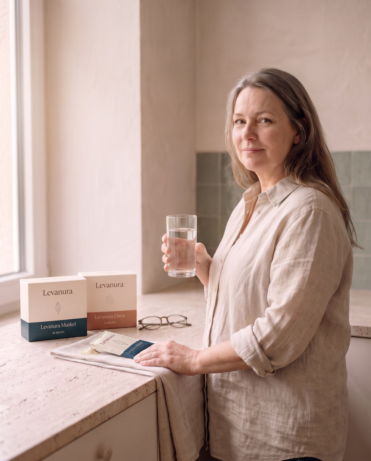 Frau in der Küche mit Glas Wasser in der Hand und Levanura Muskel und Levanura Darm Nahrungsergänzungsmittel auf dem Tresen.